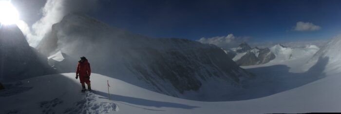 A climber on a snowy surface, looking at Everest emerging from the clouds. 