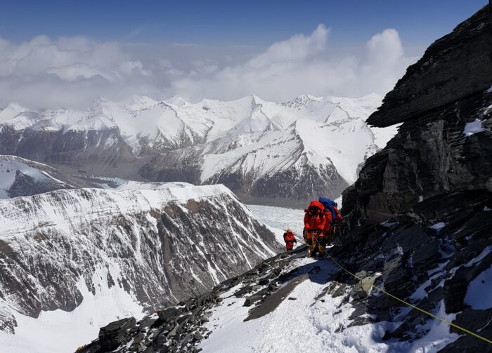Climbers at a traverse with little snow, clipped to a rope, mountains below them. 