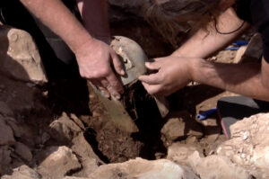 men lift a dirty gladiator-style helmet from a hole