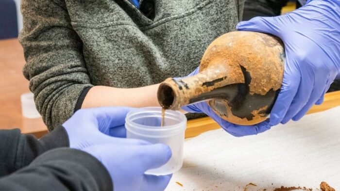 pouring liquid from a soiled glass jug into a small plastic container with gloved hands