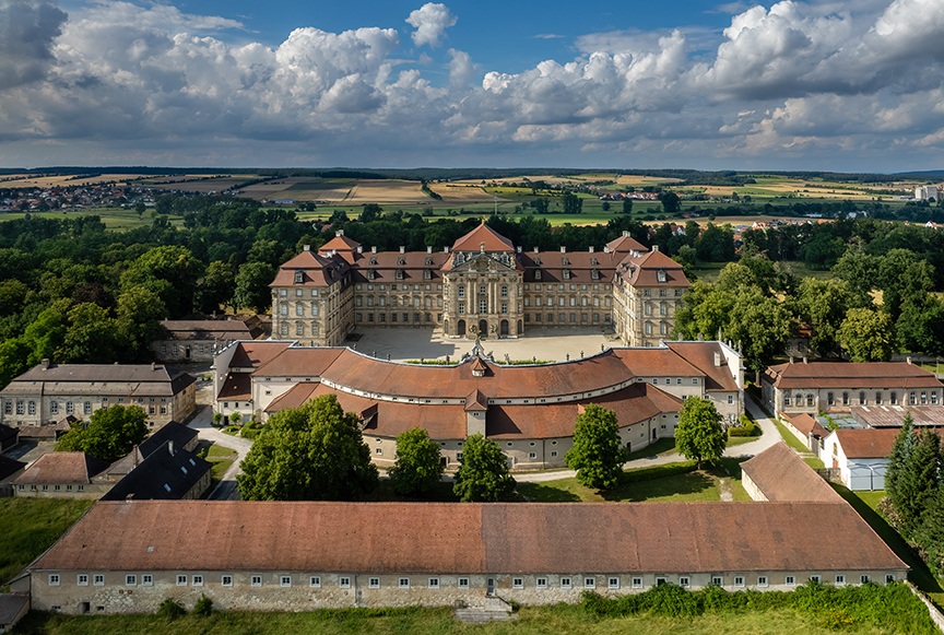 An aerial photograph of a red-roofed castle in the Baroque style, with multiple wings and a central courtyard.