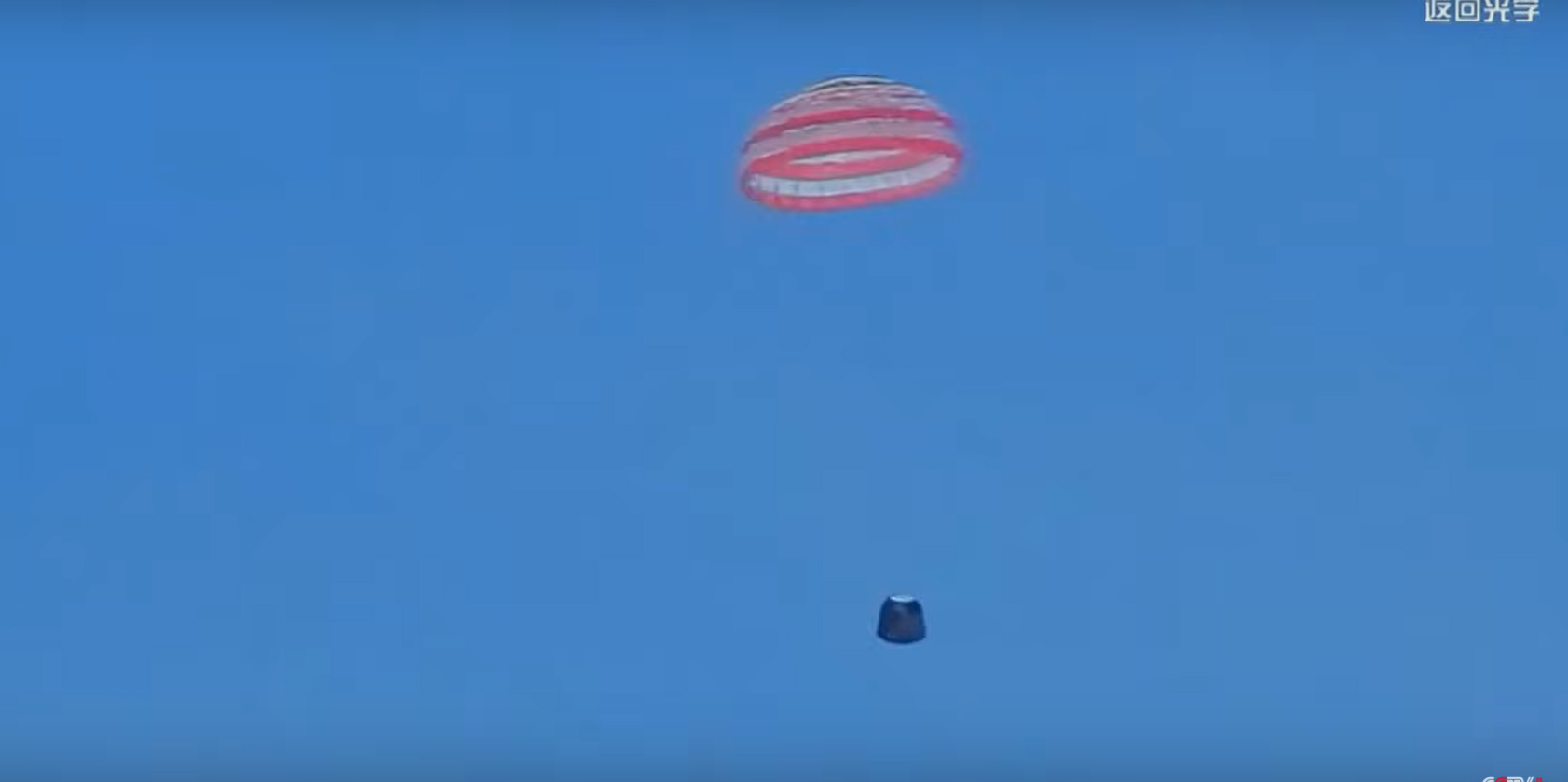 A red and white striped parachute floats through the blue sky. A dark object is suspended below it.