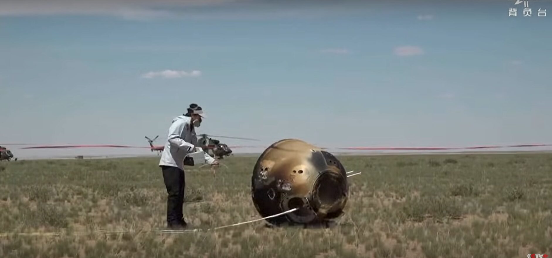 A scorched, bell-shaped container lies in a grass field. A person in a white coat holds out a Geiger counter. In the background, a helicopter waits.