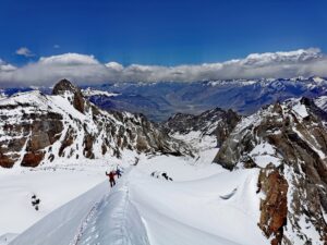 two climbers on a snowy ridge, with rock and snow peaks around.