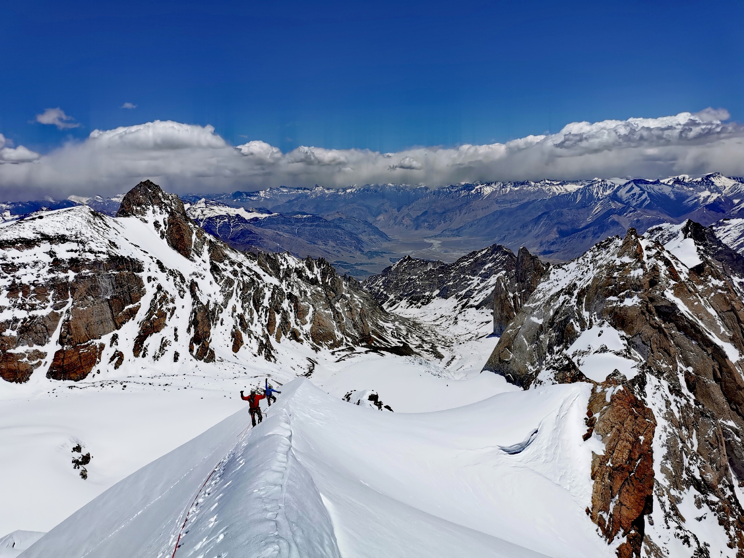 two climbers on a snowy ridge, with rock and snow peaks around.