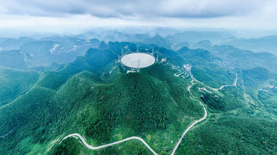A radio dish sits in a sinkhole at the top of a mountain. More mountains stretch out behind it.
