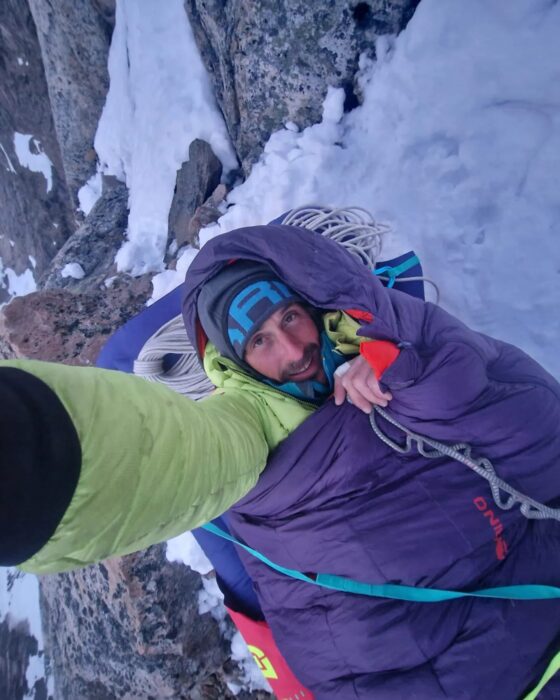 A climber takes a selfie while lying on a hammock on a big wall.