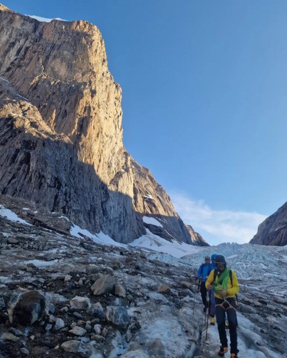 Climbers on the icy ground observe a granite face lit by the sun in Greenland.