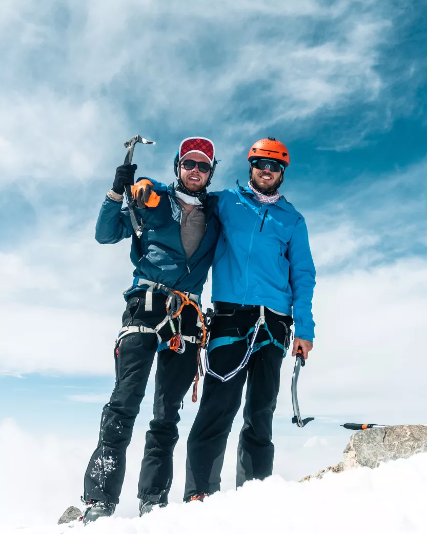 Two men stand at the top of a mountain holding ice axes.