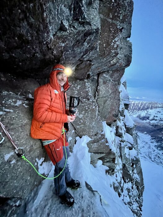 Gietl with a stove in his hand while stending on a very narrow snow ledge.