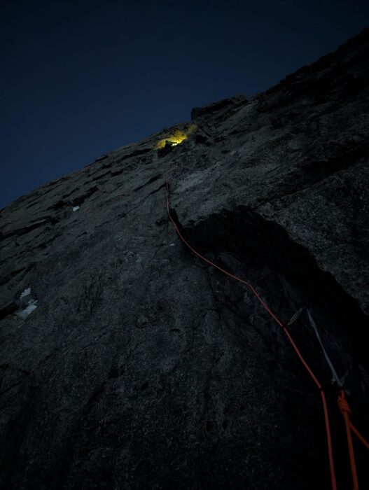 A granite face in the dark, with only the headlamp of a climber visible on the vertical rock. 