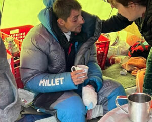 French climber with bruised face and a hand in bandages, holds a mug and speaks to a doctor in a kitchen thent at Annapurna Base Camp