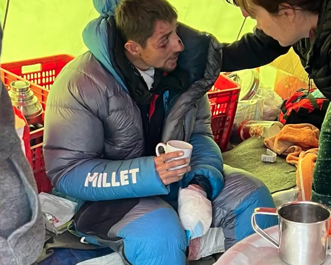 French climber with bruised face and a hand in bandages, holds a mug and speaks to a doctor in a kitchen thent at Annapurna Base Camp