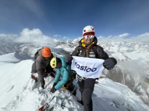 Three climbers on a snowy summit, one of them standing up with a banner.
