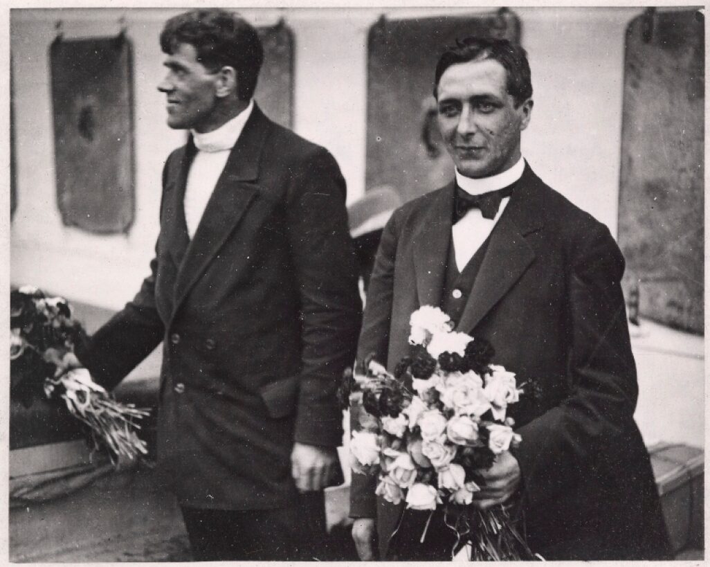 Two men in formal dress, holding flowers.