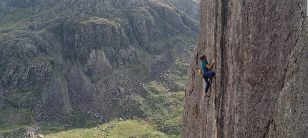 Woman climbing a sheer rockface, with a green vale in the background.