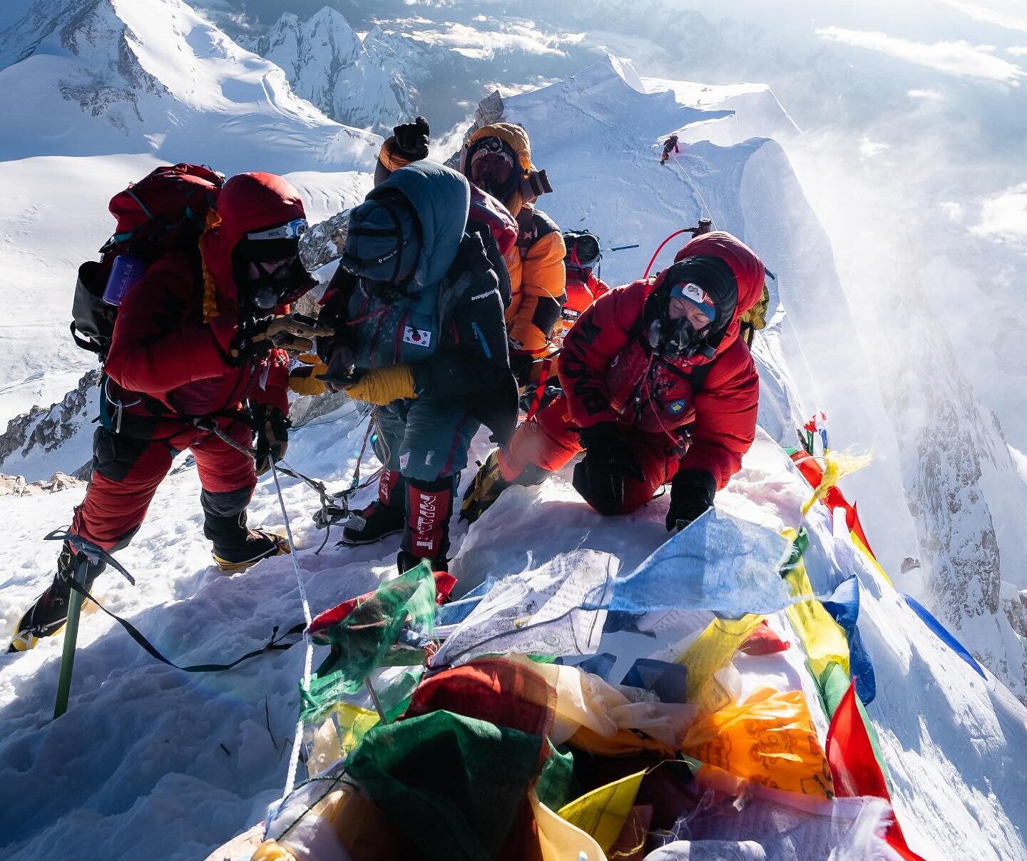 A group of sherpas by a bunch of colorful flags on the top of Makalu, the snowy ridge behind them.