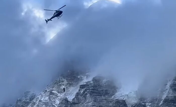 A helicopter among the clouds covering Everest flanks, carries a climber hanging at the end of a cable. 