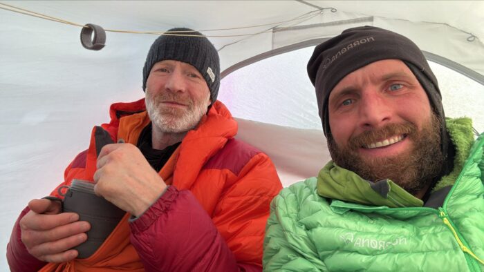 Two men wearing winter jackets and beanies smile while sitting inside a tent. Borge Ousland in a red and orange jacket holds a camping cup, and the other Vincent Colliard in a green jacket is taking the selfie. They appear to be in good spirits.