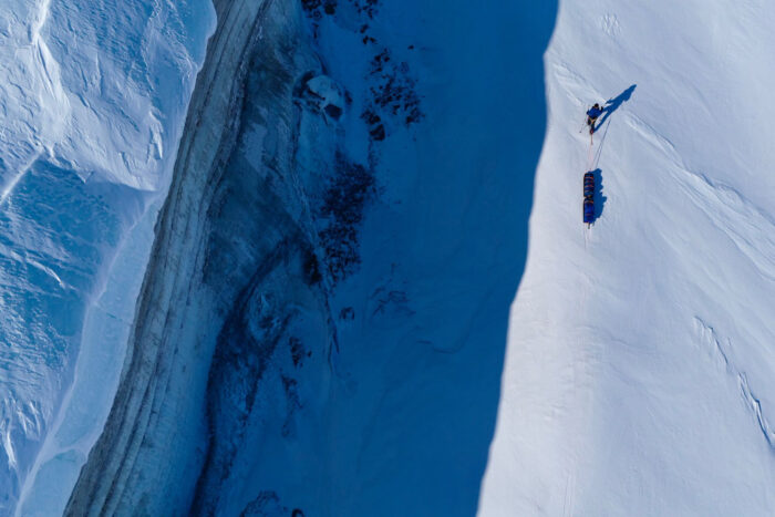 A dramatic aerial view of a lone skier - Borge Ousland - pulling a sled across a snowy scene. The skier is on the bright, sunlit side of the slope, while a deep, dark shadow sharply divides the scene. To the left, a steep ice wall and rugged terrain highlight the harsh High Arctic environment.