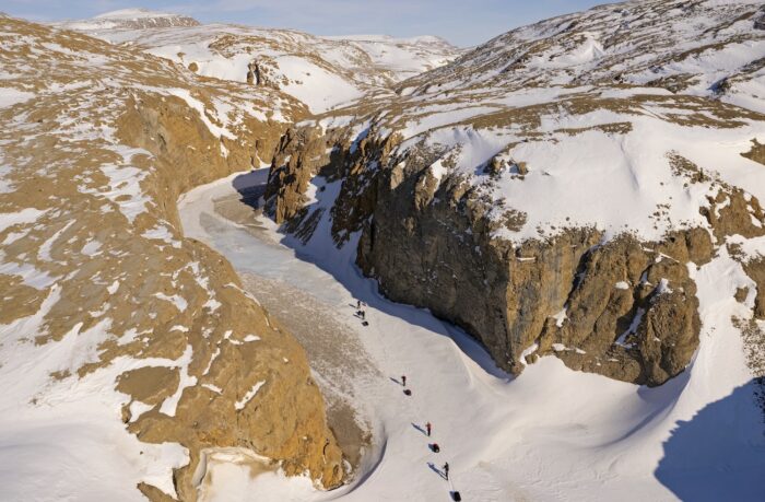 Aerial view of a narrow, snow-covered canyon winding through steep, rugged cliffs. A small group of Arctic travelers pulling sleds is visible on the frozen ground, emphasizing the scale and remoteness of the landscape. Patches of snow cling to the brown rock formations, and the frozen riverbed below forms a natural path through the terrain. The image captures the harsh, wind-swept conditions of Sverdrup Pass.