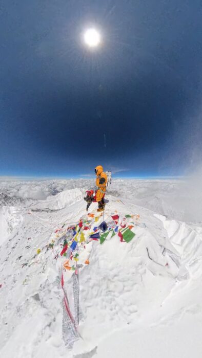 A climber on the summit of Everest, wind rising from the flank and prayer flags on the snow. 
