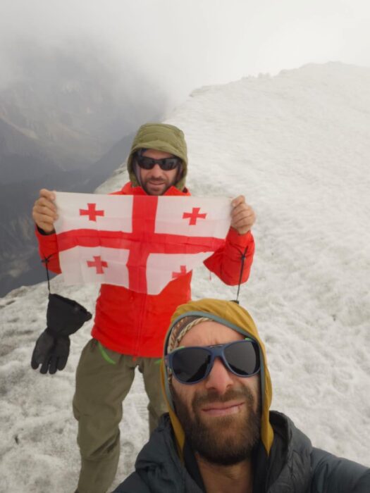 Two climbers hold a Georgian flag on a misty summit.