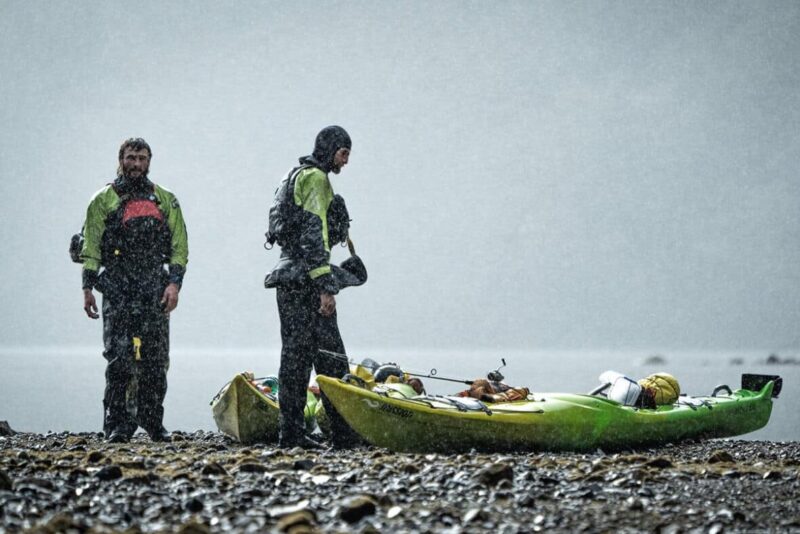 Two kayakers stand next to their loaded boats in the pouring rain