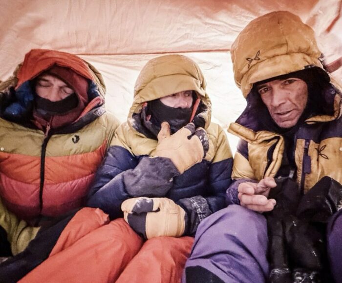Three climbers looking cold and tired with all their clothes on, in a tent. 