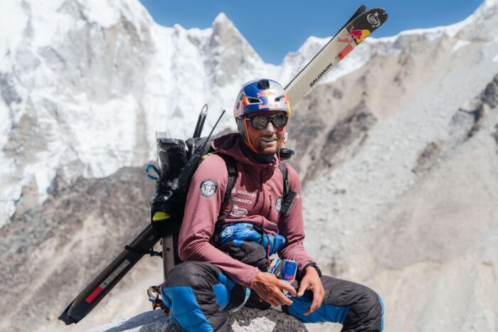Bargiel in a tshirt and helmet, with skis in backpack, sitting on a rock.