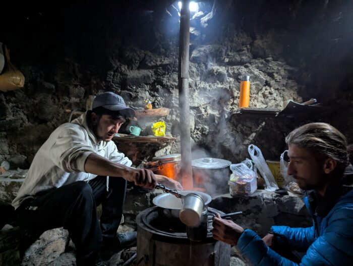 Two climbers cook at a small fire in a stone cabin. 