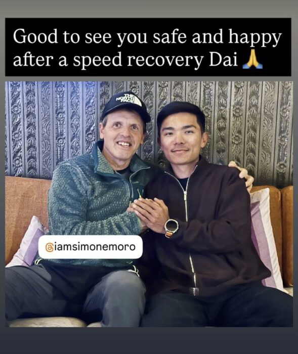 Simone Moro and Nima Rinji Sherpa hold hands while sitting on a coach.