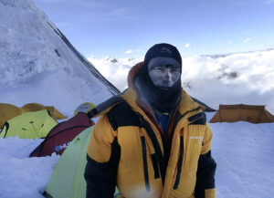 Sanu Sherpa on a snowy plateau with some high altitude tents behind him.