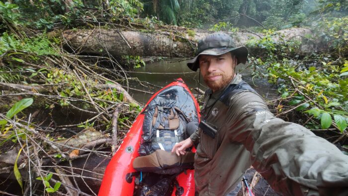 Swiss adventurer Loic Cappellin poses next to his red packraft on a river clogged with fallen trees