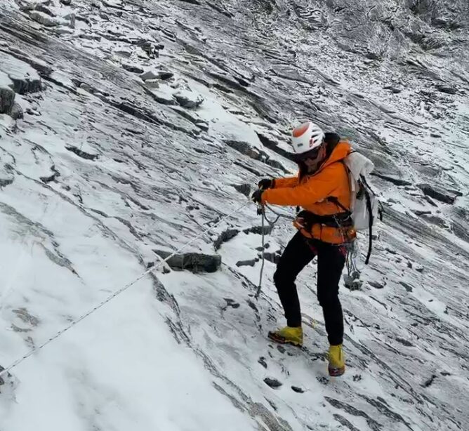 A climber clipped to afized rope descends on a slippery slope of scree covered with some fresh snow.