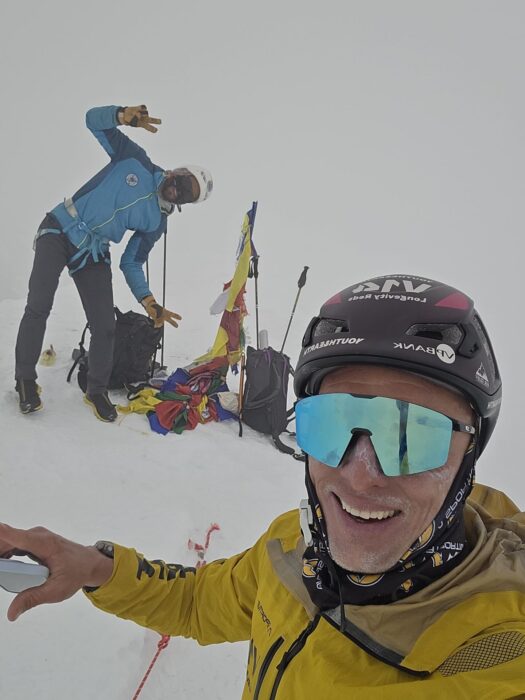 Climbers greet to the camera from a snowy summit in a very foggy day.