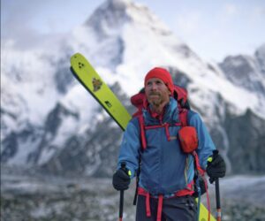 A climber walking on morraine terrain with skis on his backpack and a peak behind him.