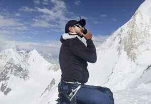Close shot of Purja speaking on the radio from a snowy slope, peaks in background.