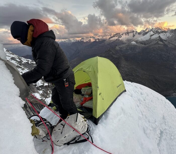 A climber belaying himself to a mixed face while he has set a tiny tent behind him on a snow mushoom.