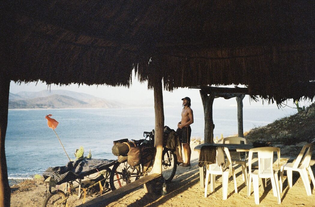 Tristan La Hayes stands on a beach with his bike and surfboard